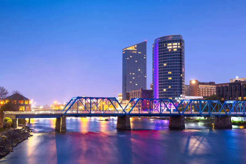Downtown Grand Rapids skyline at dusk with the Grand River and a lit bridge in the foreground
