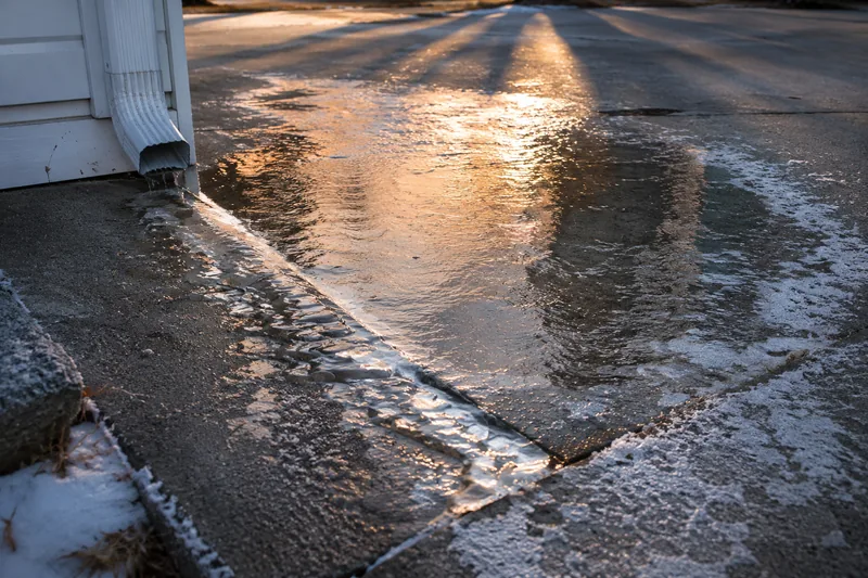 Downspout discharging across concrete slabs in low winter sun, the runoff refrozen into a sheet of ice over the pavement