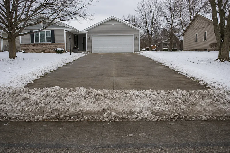 Compacted wall of grey plow snow left across the bottom of a cleared residential driveway where it meets the street