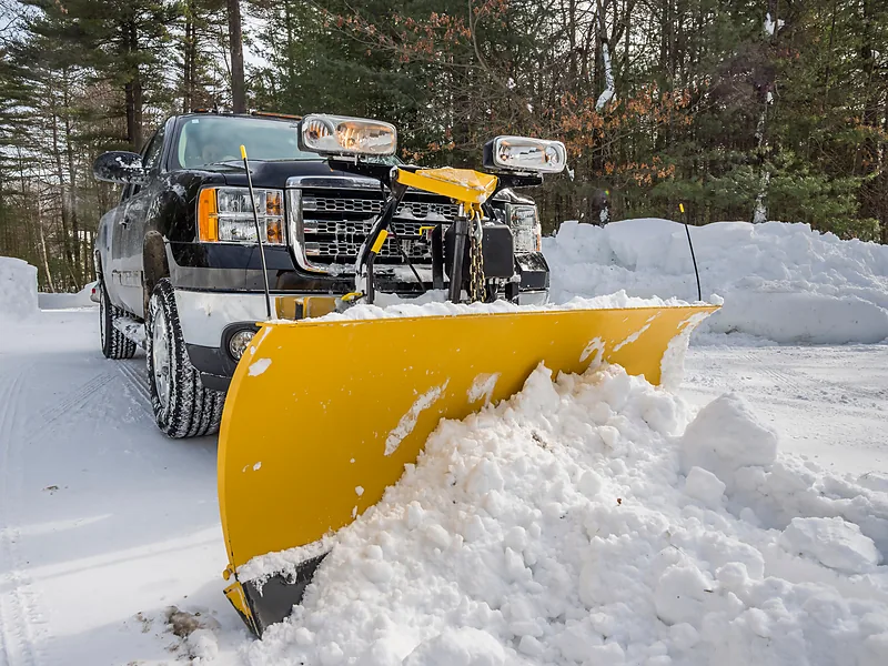 Pickup-mounted plow blade angled into deep packed snow, marker rods set on the blade corners