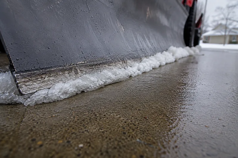 Ground-level close-up of a plow blade cutting edge scraping a driveway down to bare wet concrete, a ribbon of packed snow curling along it