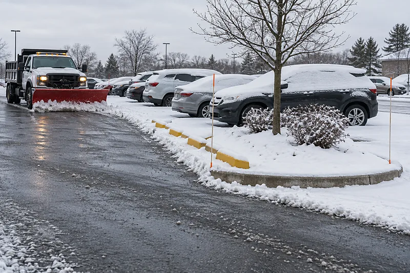 Plow truck clearing a lane past a row of snow-covered parked cars, with a landscaped island, yellow curb stops and orange-tipped marker stakes alongside