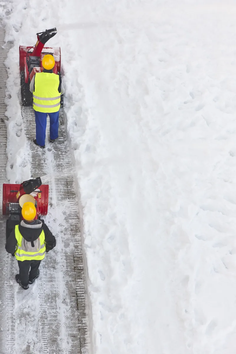 Overhead view of two crew members running snow blowers single file along a sidewalk, the cleared full-width strip set against untouched deep snow beside it