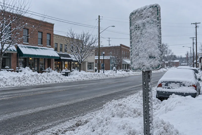 Snow-crusted street sign on a post beside a plowed main street, snow-covered cars at the curb and low brick storefronts beyond