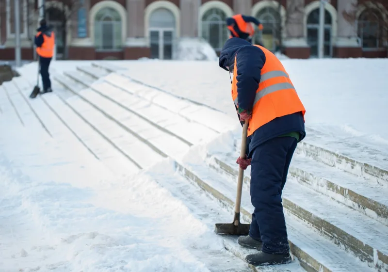 Two crew members in high-visibility vests clearing snow by hand from the wide entrance steps of a building