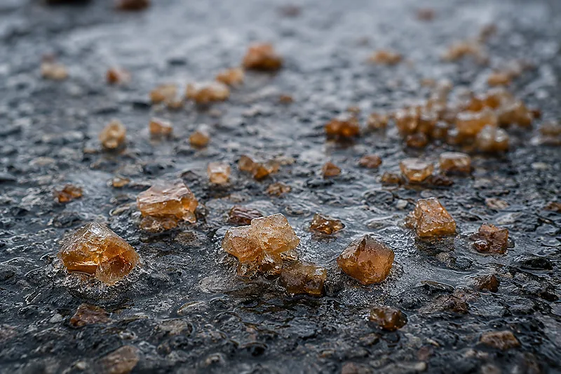 Close-up of amber-tinted treated rock salt granules pre-wetted with brine, scattered across wet dark asphalt with meltwater haloes forming around them