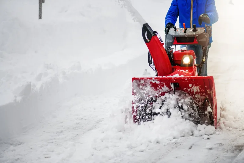 Operator pushing a walk-behind snow blower through deep snow, chute throwing a plume to the side
